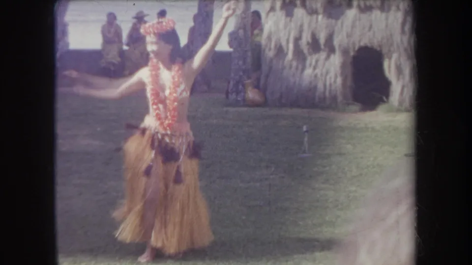 1969 Hawaii home-movie frame showing a hula dancer in lei, headpiece, and grass skirt gesturing on a lawn before onlookers.