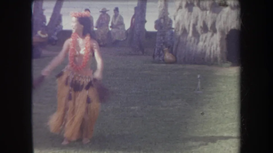 1969 Hawaii 8mm still of a hula dancer moving in a grass skirt and floral lei, with people seated near palms and a thatched backdrop.