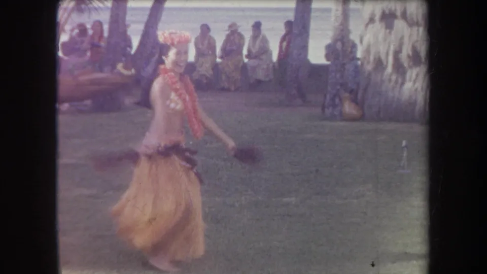 1969 Hawaii home-movie frame of a hula dancer turning with arms extended, filmed on a lawn with spectators, palms, and ocean beyond.