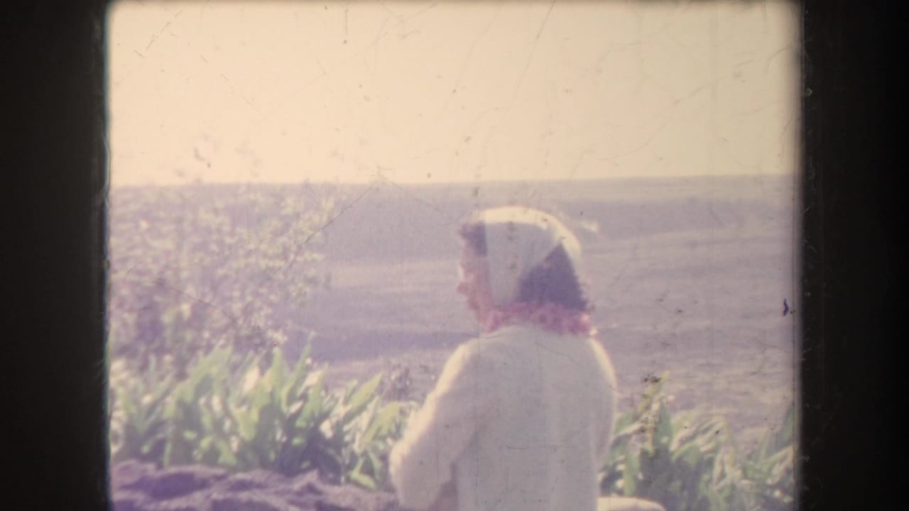 1969 Hawaii home-movie frame showing a woman in light clothing and headscarf beside coastal vegetation with the ocean in the background.
