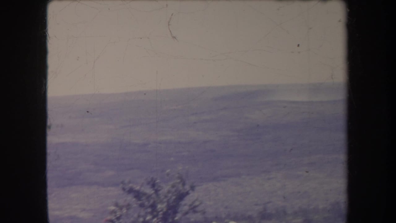 1969 8mm frame with a hazy distant ridge line, pale sky, dark film borders, and a small shrub shape near the lower edge.