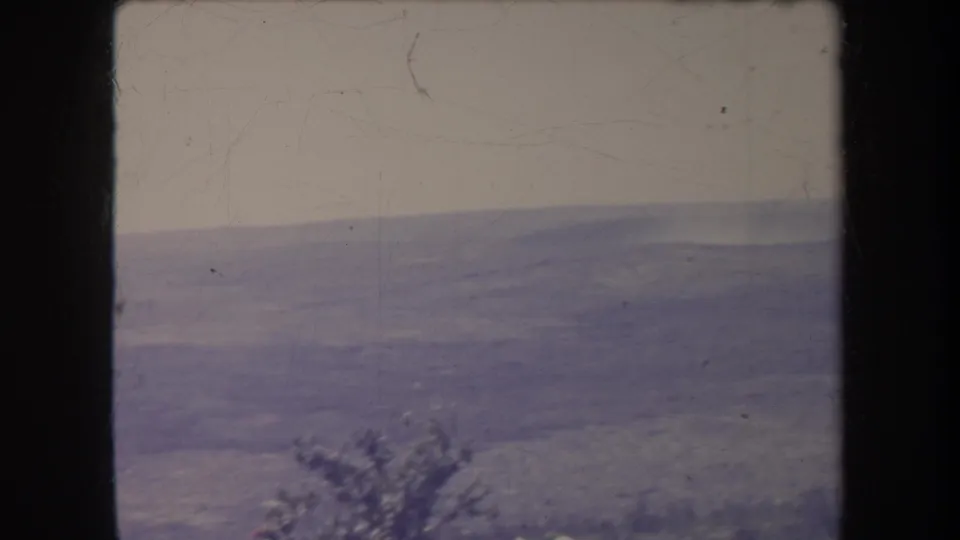 1969 8mm frame with a hazy distant ridge line, pale sky, dark film borders, and a small shrub shape near the lower edge.