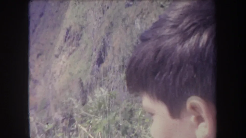 Young boy, likely in a 1969 Hawaii home movie, looks downward near rocky dirt and sparse vegetation.