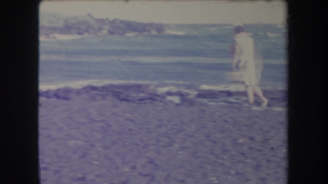 1969 Hawaii 8mm frame showing a woman in light clothing walking near the water on a stony beach with surf behind her.