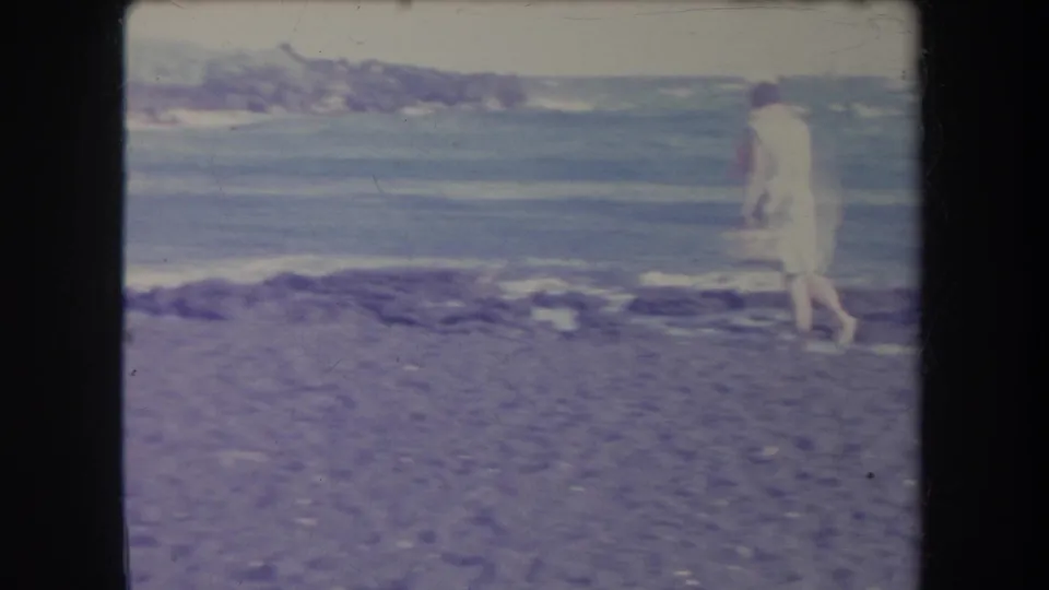 1969 Hawaii 8mm frame showing a woman in light clothing walking near the water on a stony beach with surf behind her.