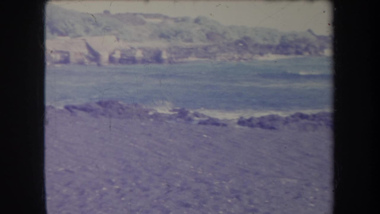 Rocky beach and water with distant shoreline buildings and green hills.