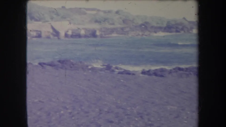 1969 Hawaii coastal view with a stony foreground, calm water, distant buildings, and green hills in faded 8mm color.