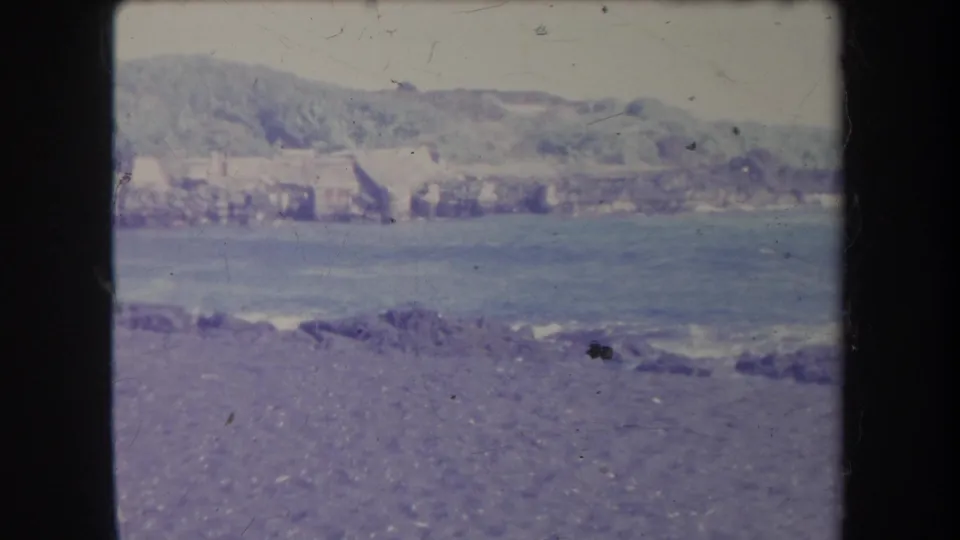 1969 Hawaii shoreline frame showing stones in the foreground, water beyond, and a distant hillside settlement across the bay or inlet.