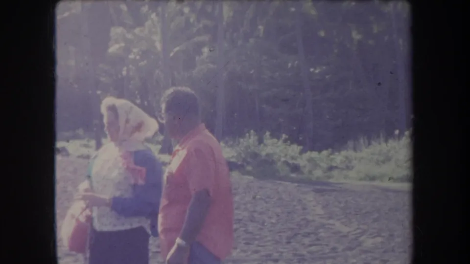 1969 Hawaii home-movie frame showing two adults on a rocky beach, one with a head covering and bag, near shaded coastal vegetation.