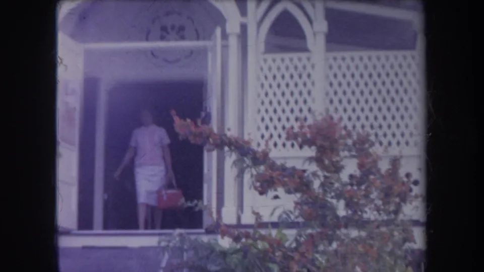 1969 Hawaii home-movie frame showing an adult at a bungalow doorway, with white lattice siding and flowering plants nearby.
