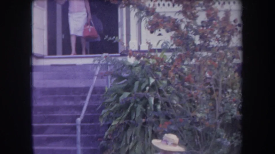 1969 Hawaii 8mm frame showing a woman near house steps, white lattice siding, and dense tropical garden foliage.
