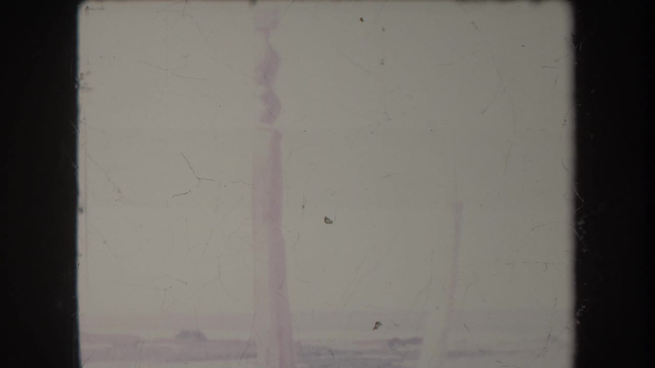 1969 Hawaii shoreline frame with a faint surf line and distant vertical structure, softened by color fading and dust.