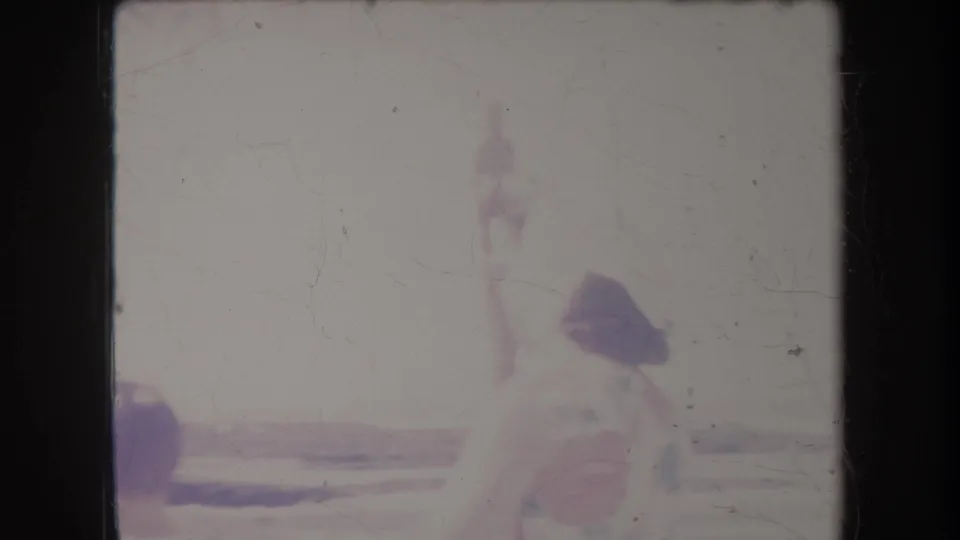 1969 Hawaii home-movie frame with a dark beach umbrella shape and faint human figure near the shoreline amid strong color fading.