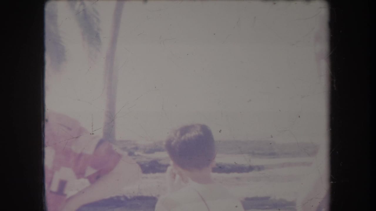 1969 Hawaii 8mm frame showing people from behind at a very bright outdoor gathering near palm trees and open coastal light.