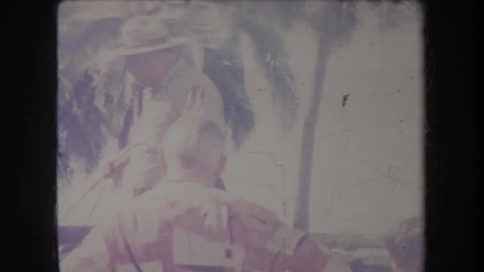 1969 Hawaii home-movie frame with adults gathered in harsh sunlight beneath palms, partly washed out by exposure.