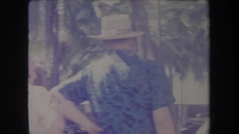 1969 Hawaii 8mm frame showing a man in a light hat and patterned shirt at an outdoor picnic or gathering with palms behind him.