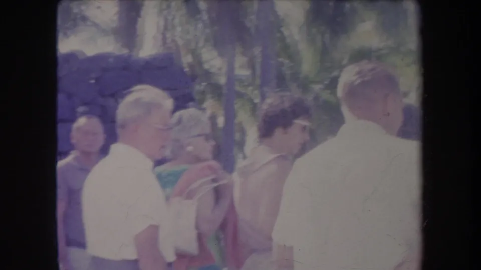 1969 Hawaii frame showing adults in light summer clothing gathered near palm trees and a dark stone wall in bright daylight.