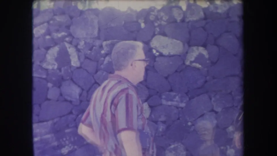 A man in glasses and a striped shirt passes in front of a dark stone wall in 1969 Hawaii home-movie footage.
