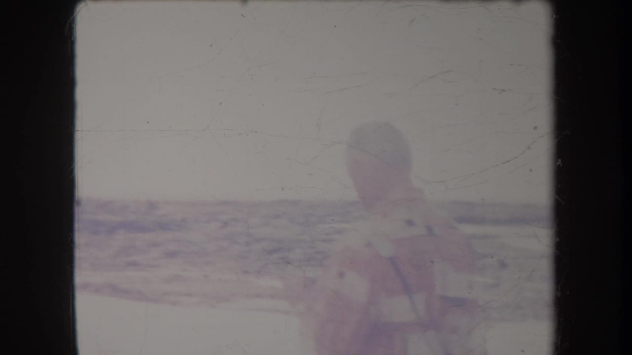 Older man holding binoculars beside a bright tropical beach and waterline.