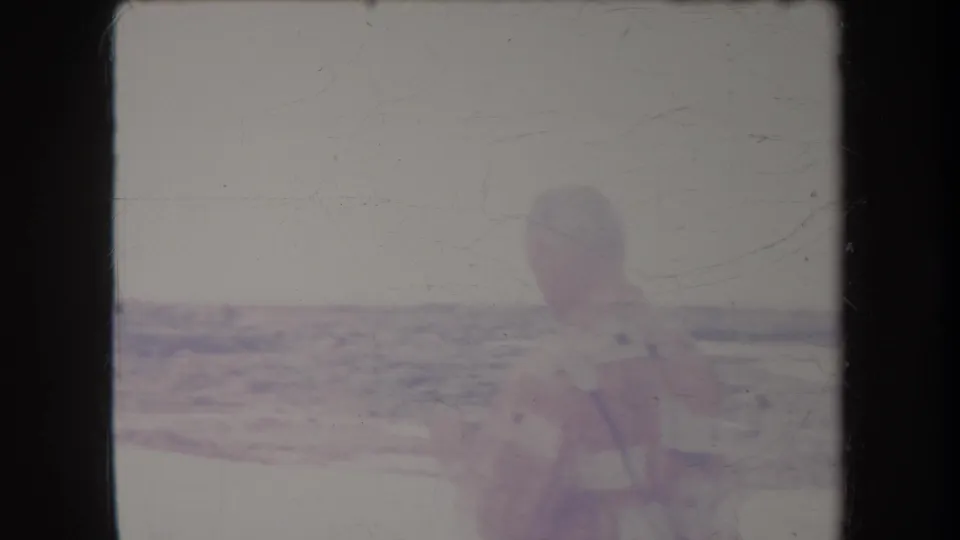 1969 Hawaii home-movie frame with an older man using binoculars near a bright beach, water, and distant shoreline.
