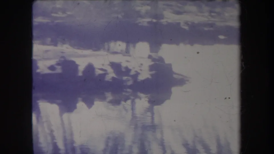 1969 home-movie frame showing muted swamp reflections, shoreline darkness, and vertical tree shapes in a purple-shifted scan.