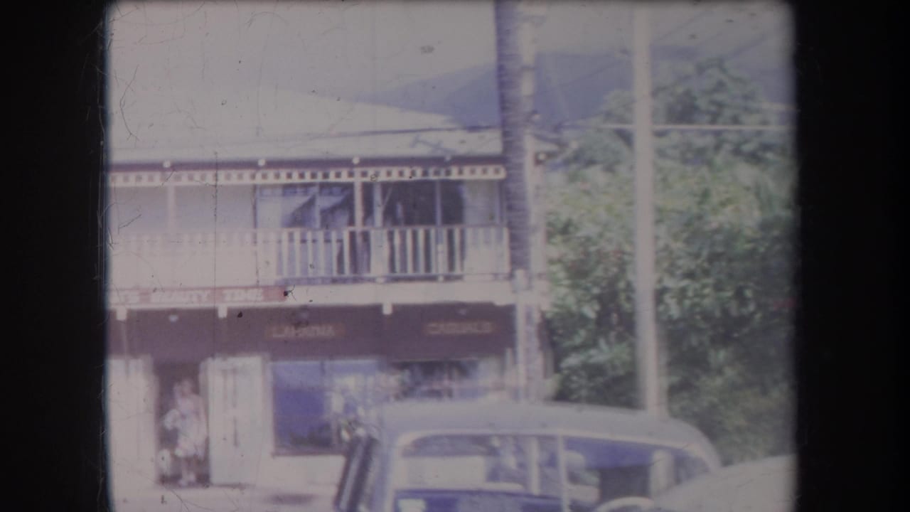 1969 Hawaii home-movie frame showing a two-story Lahaina-area storefront, balcony railing, parked vintage car, and a pedestrian.
