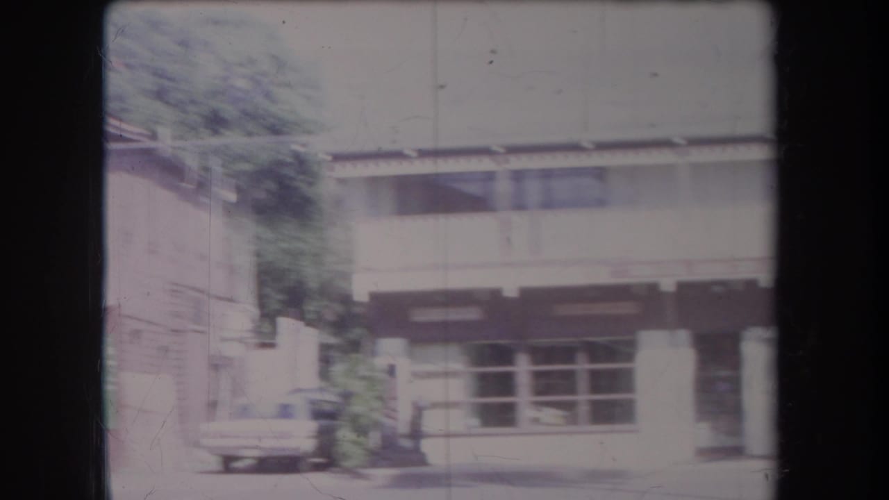 Light-colored building with broad windows, posts, greenery, and a parked car near the curb.