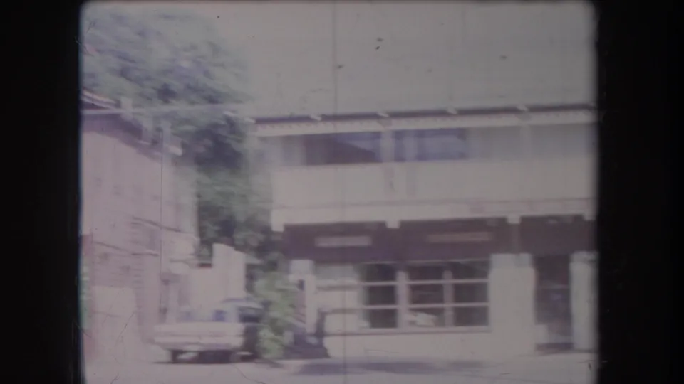 1969 Hawaii street view with a low storefront facade, shaded windows, sidewalk posts, vegetation, and a parked vintage car.