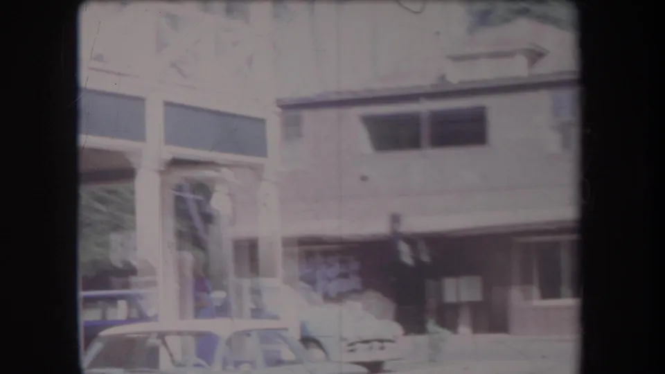 Faded 1969 Hawaii home-movie frame showing older storefront buildings, multiple parked cars, and a traffic signal on a town street.
