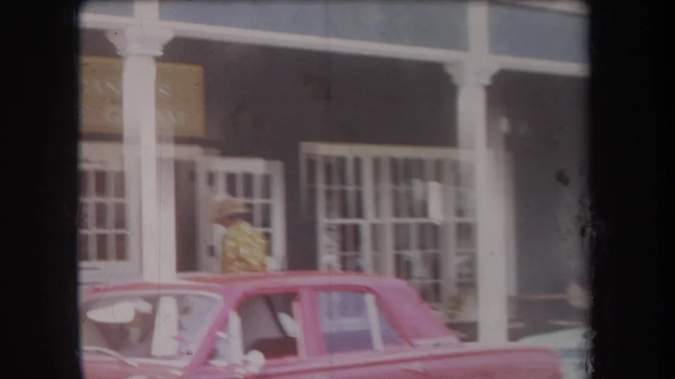 1969 Hawaii street frame with a pink vintage car, white storefront posts, barred windows, and a person in a straw hat passing by.