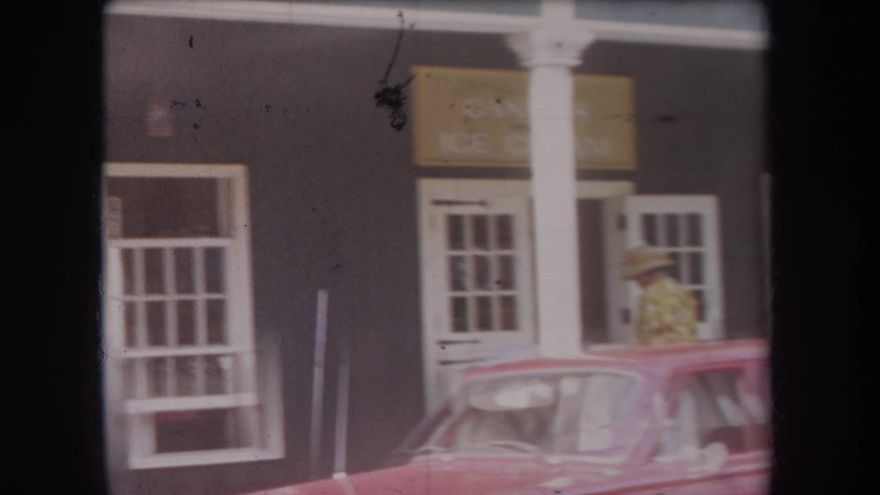 1969 Hawaii home-movie frame showing a dark storefront, white trim, red car, and a person in a straw hat near the entrance.