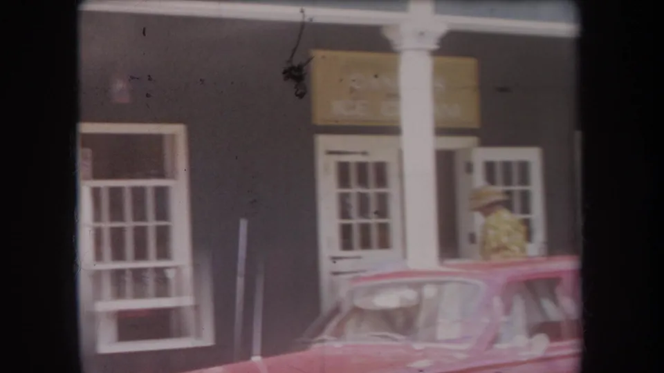 1969 Hawaii home-movie frame showing a dark storefront, white trim, red car, and a person in a straw hat near the entrance.