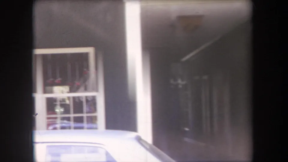 1969 Hawaii home-movie frame showing a storefront window, pale car roof, and dark hallway-like interior in soft exposure.