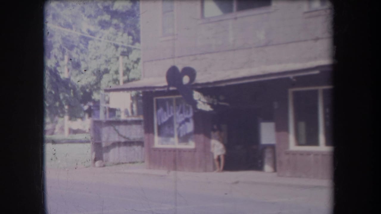 1969 Hawaii home-movie frame showing a shaded storefront with painted signage and a woman in a light dress near the doorway.