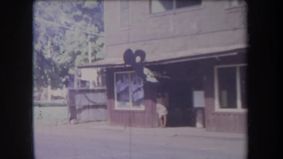 1969 Hawaii home-movie frame showing a shaded storefront with painted signage and a woman in a light dress near the doorway.