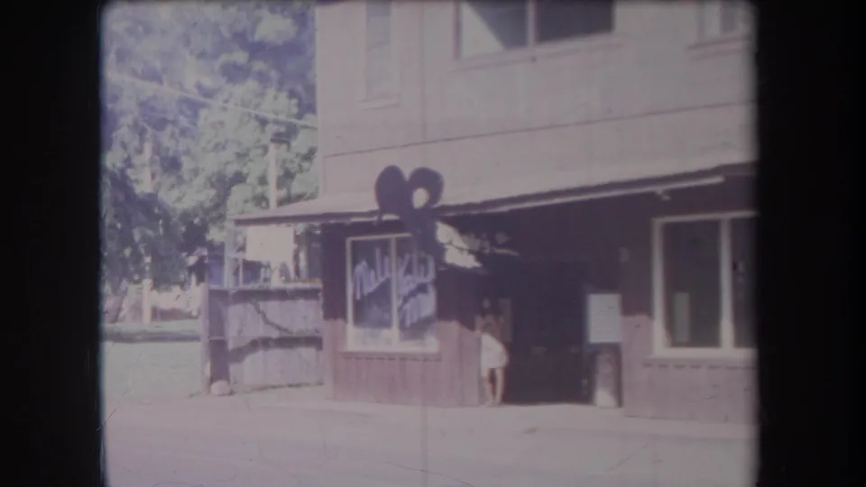 1969 Hawaii street view of a small shop facade, painted sign, shaded entrance, and woman standing by the doorway.