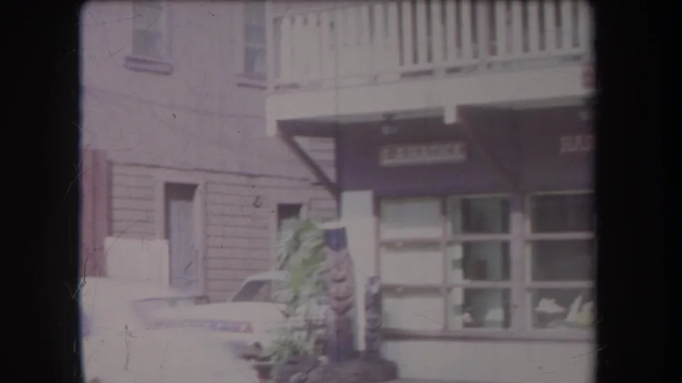 1969 Hawaii home-movie frame showing a commercial facade with balcony, signboard, parked classic car, and tiki-style figure near the sidewalk.