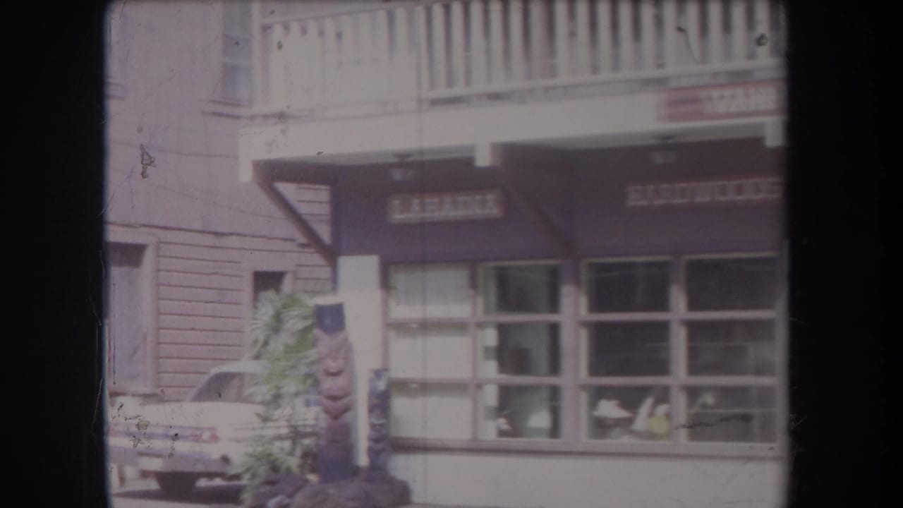 1969 Hawaii home-movie frame showing a storefront block with awnings, large display windows, a parked car, and partially readable shop signs.