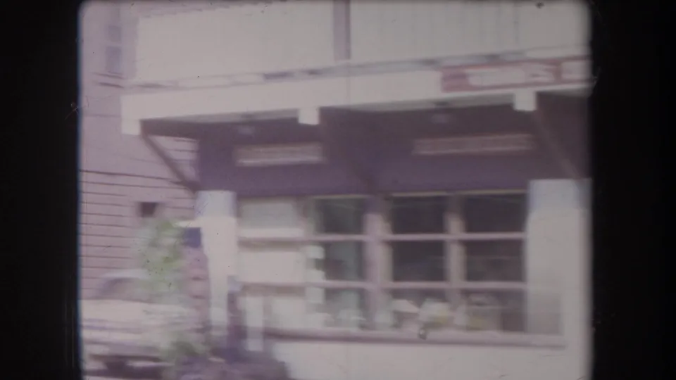 Soft 8mm frame of a Hawaii downtown storefront in 1969, with awnings, display windows, a parked car, and partial signage visible.