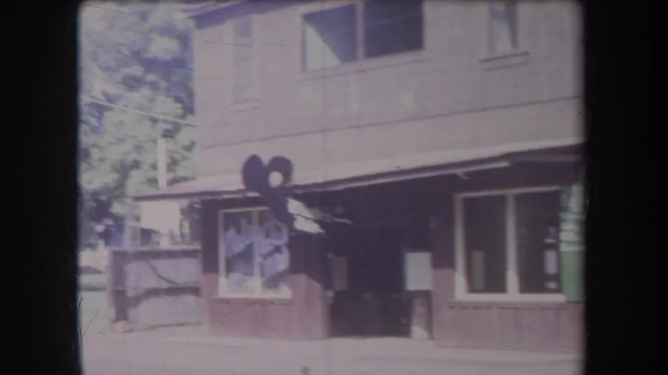 1969 Hawaii home-movie frame showing a commercial building exterior with a shaded doorway, large window, and tiki-style figure outside.