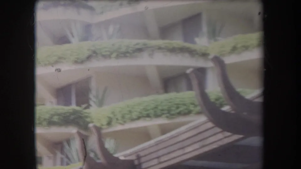 1969 Hawaii 8mm frame of a tropical exterior with planted ledges, balcony recesses, and dark curved beams in the foreground.