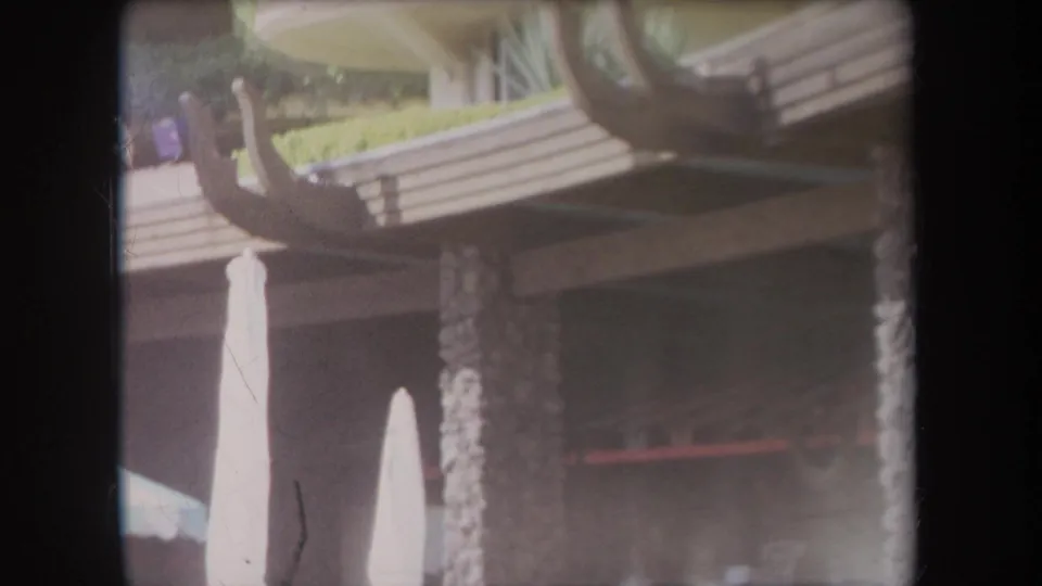 1969 Hawaii 8mm frame showing an exterior with umbrellas, a palm trunk, planted concrete ledges, and curved wooden beams.