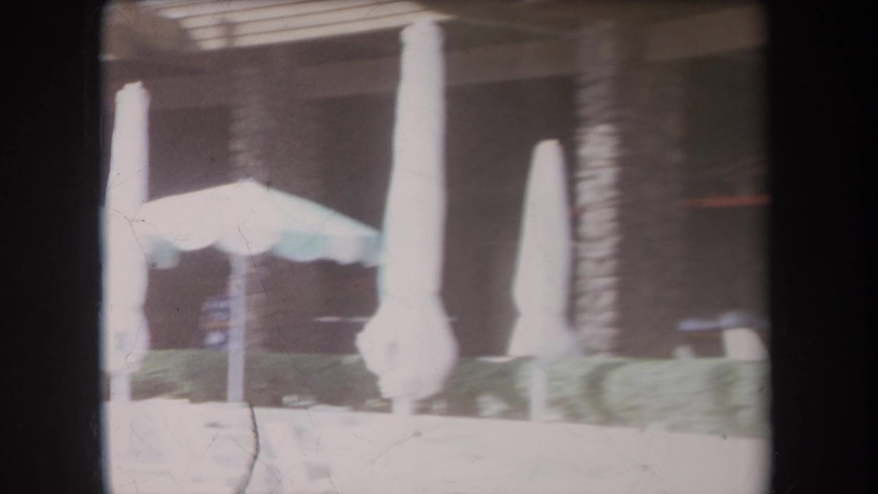 1969 Hawaii resort patio with closed umbrellas, white lounge seating, hedges, palm trunks, and stone columns in soft 8mm exposure.