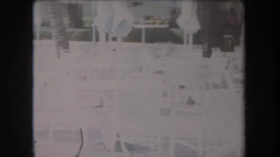 Bright 1969 Hawaii resort patio view with white chairs, tables, closed umbrellas, hedges, and palm trunks softened by 8mm exposure.