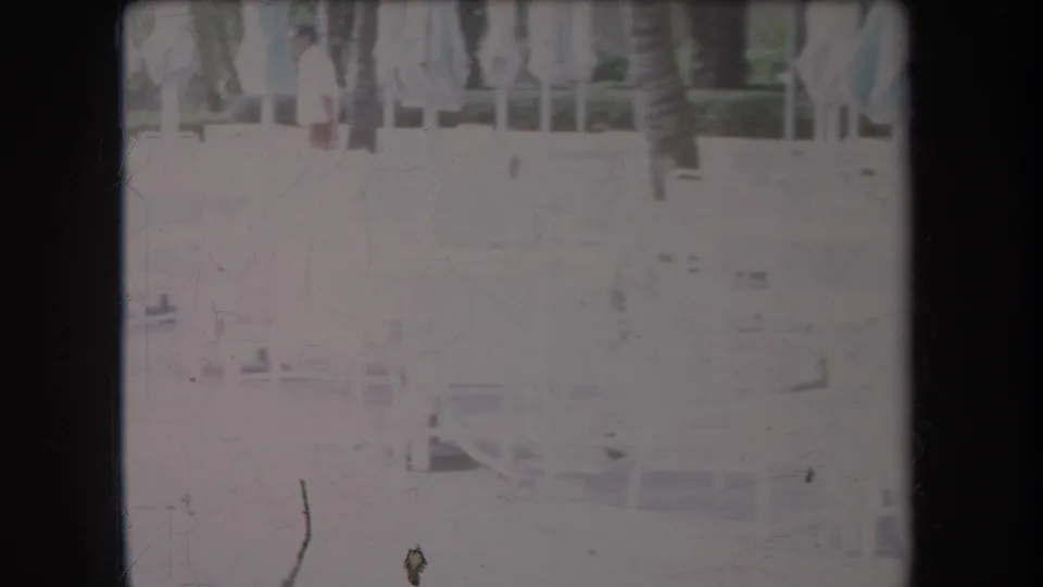 A distant person crosses a 1969 Hawaii resort patio behind white chairs, tables, closed umbrellas, and palm trunks in faded 8mm footage.