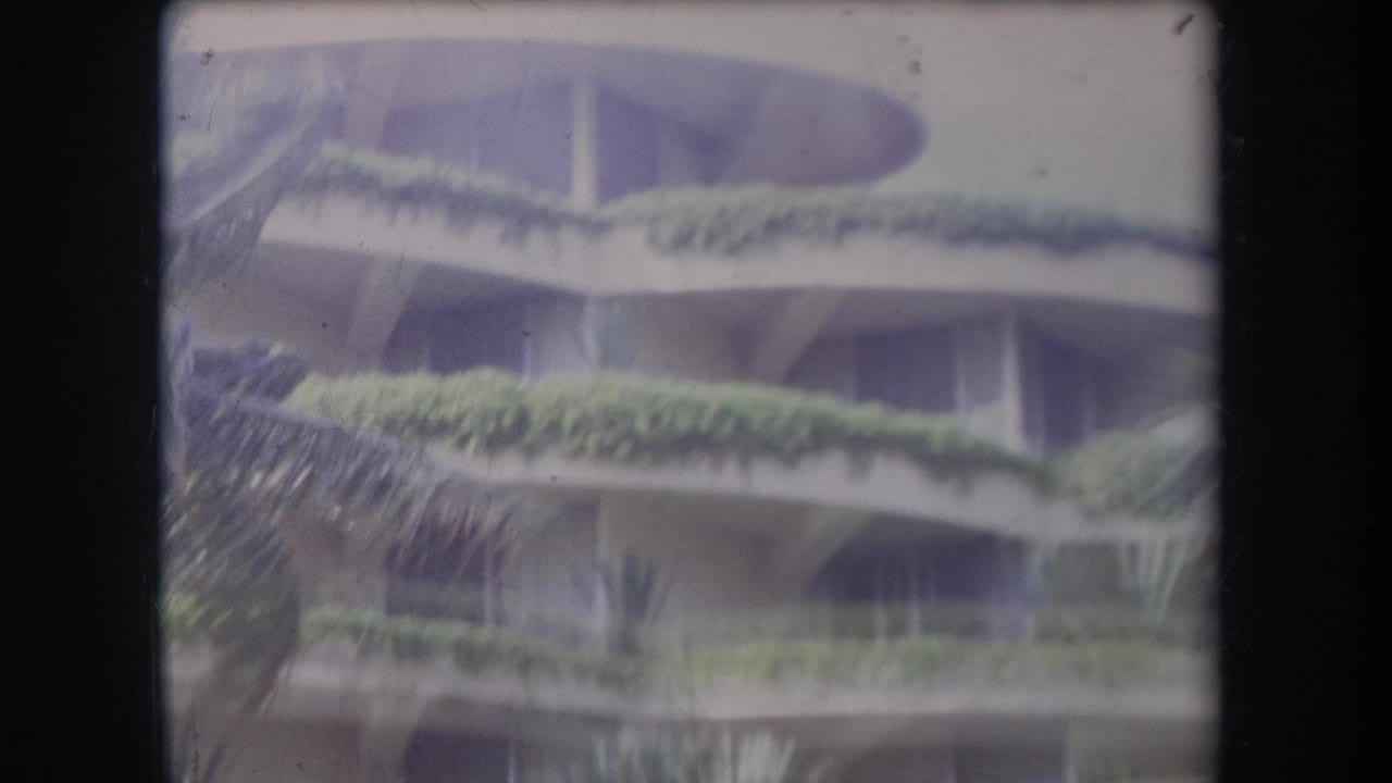 Multi-story building with planted balconies seen through blurred palm movement.