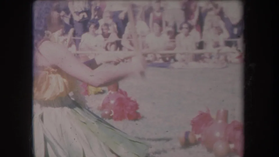 1969 Hawaii 8mm frame showing two women in hula-style costumes dancing before spectators behind a barrier.