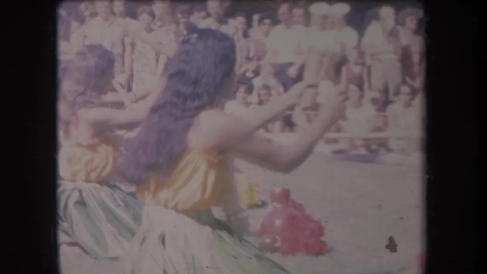1969 Hawaii home-movie frame with two women in bright dance costumes performing in front of a seated and standing crowd.