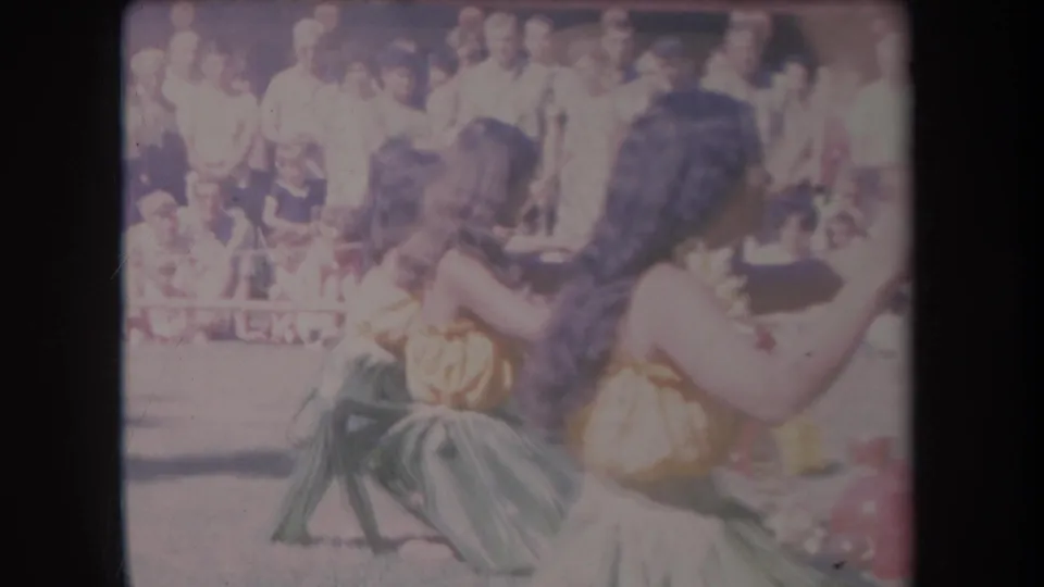 1969 Hawaii 8mm frame showing two costumed women mid-dance, with faces of spectators visible behind them.