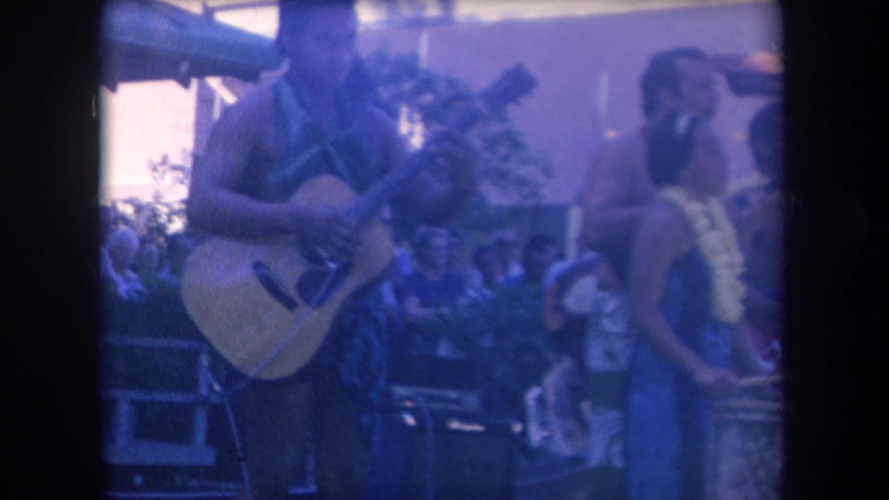 Guitar player performing on an outdoor stage with dancers and audience nearby.